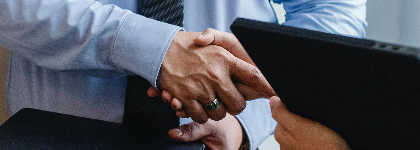 two business workers shaking hands while holding laptops